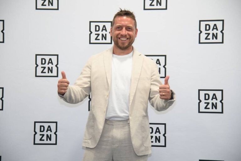Emanuele Giaccherini posa sorridente davanti al backdrop DAZN, vestito in abito chiaro durante un evento ufficiale dell’emittente sportiva.