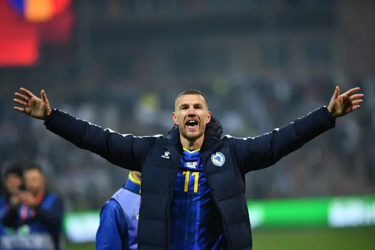 Edin Dzeko esulta con le braccia alzate indossando la maglia della Bosnia, stadio sullo sfondo