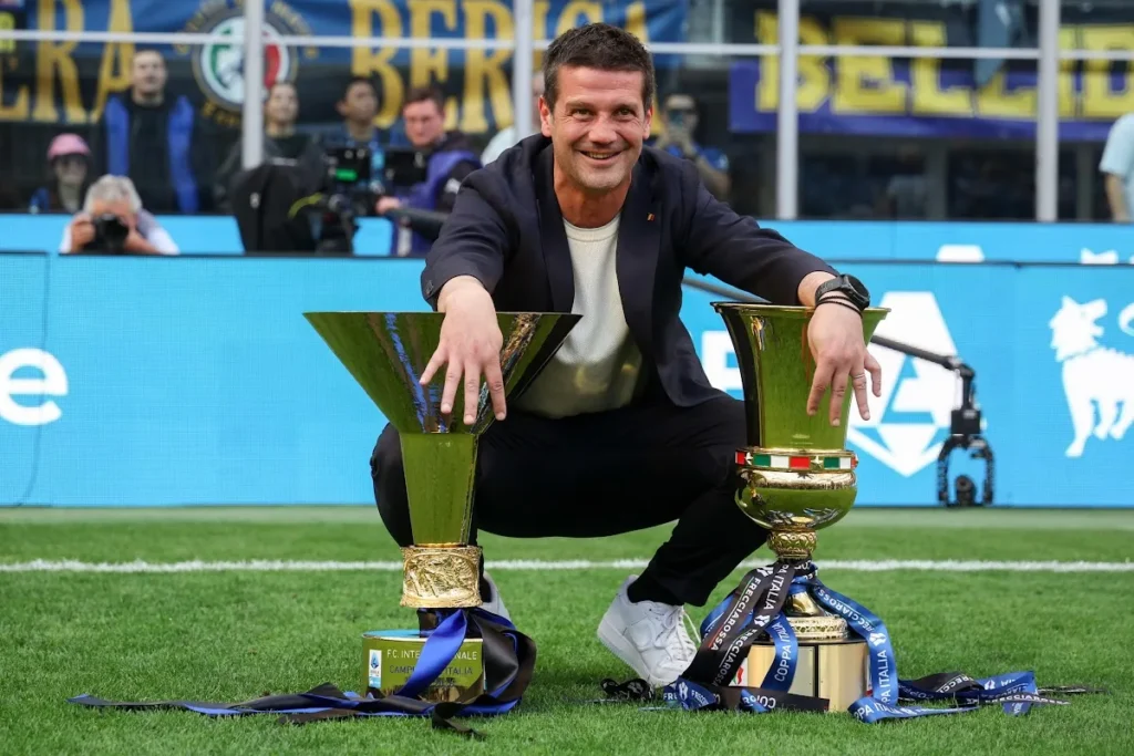 Chivu sorride mostrando due trofei sul campo dell'Inter, con lo stemma nerazzurro sullo sfondo