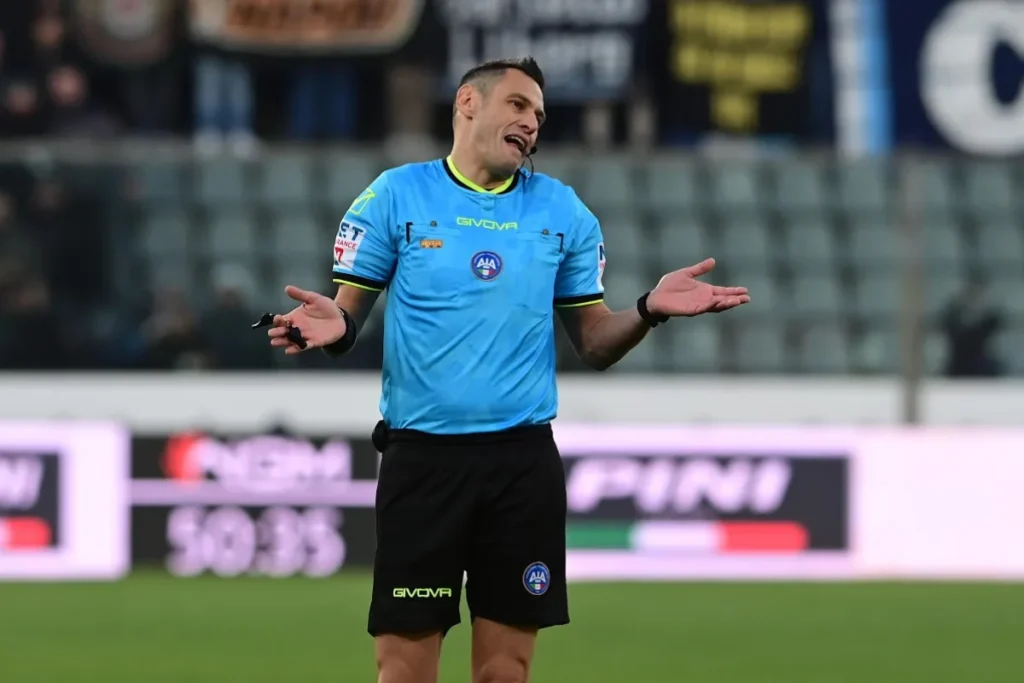 Arbitro con divisa azzurra e pantaloncini neri gesticola con le braccia aperte durante una partita di Serie A